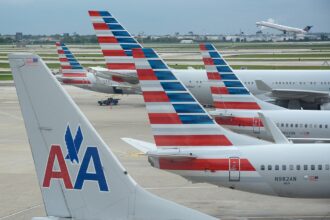 American Airlines aircraft at O'Hare that are parked side-by-side causing the brightly coloured aircraft tails to overlap each other. An aircraft is taking off in the distance into a clear sky.