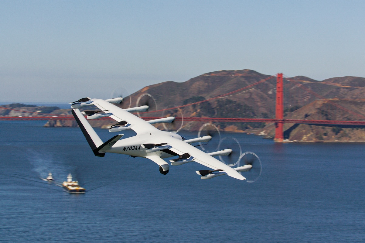 An electric air taxi with multiple propellers flies over the San Francisco Bay near the Golden Gate Bridge on a clear sunny day.