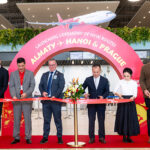 Six people stand in a row holding scissors to cut a red ribbon printed with Vietjet branding, in an airport check-in hall. Behind them is a large pink arch reading “Launching ceremony of new routes, Almaty, Hanoi & Prague,” with a graphic of a red and white Vietjet aircraft above it.