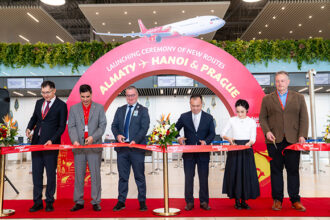 Six people stand in a row holding scissors to cut a red ribbon printed with Vietjet branding, in an airport check-in hall. Behind them is a large pink arch reading “Launching ceremony of new routes, Almaty, Hanoi & Prague,” with a graphic of a red and white Vietjet aircraft above it.