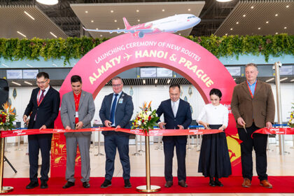 Six people stand in a row holding scissors to cut a red ribbon printed with Vietjet branding, in an airport check-in hall. Behind them is a large pink arch reading “Launching ceremony of new routes, Almaty, Hanoi & Prague,” with a graphic of a red and white Vietjet aircraft above it.