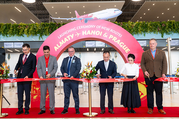 Six people stand in a row holding scissors to cut a red ribbon printed with Vietjet branding, in an airport check-in hall. Behind them is a large pink arch reading “Launching ceremony of new routes, Almaty, Hanoi & Prague,” with a graphic of a red and white Vietjet aircraft above it.