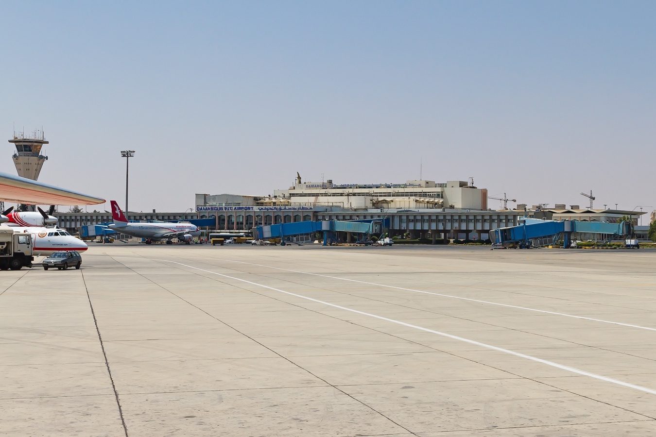 Damascus International Airport terminal with blue jet bridges, parked aircraft and a control tower beside a wide, empty apron.