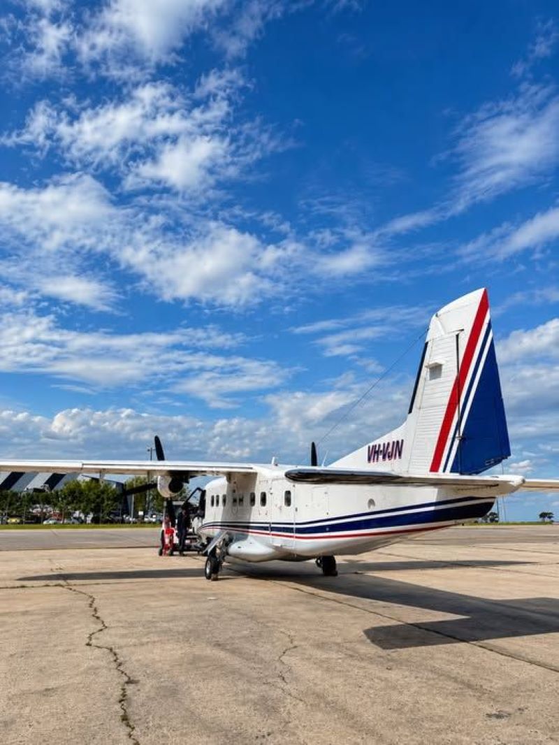 A Dornier 228-200 photographed from the back of the aircraft, with its fuselage facing forward. The aircraft has a small design on its tail - it is striped with navy, white and red. The plane is stationed on a tarmac surrounded by sunny weather and limited clouds.