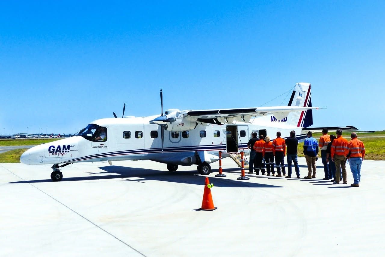 A Dornier 228 aircraft is picking up a group of people for work purposes. The men are dressed in orange jumpsuits and are boarding the plane, which is on a runway, ready to take off. The sky is bright blue and clear, with not a cloud in sight.