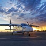 GAMair's Dornier aircraft stationed on an airport tarmac. The sun is setting in the sky, creating an ombre from blue to yellow. Under heavy cloud cover, the aircraft is distinguishable by its unusual shape - it has a pointed nose that sticks out past the cockpit and two propellers on either side of its wings.