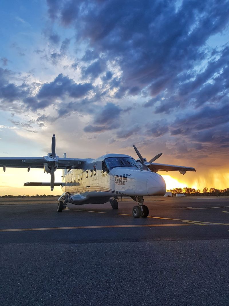 GAMair's Dornier aircraft stationed on an airport tarmac. The sun is setting in the sky, creating an ombre from blue to yellow. Under heavy cloud cover, the aircraft is distinguishable by its unusual shape - it has a pointed nose that sticks out past the cockpit and two propellers on either side of its wings.