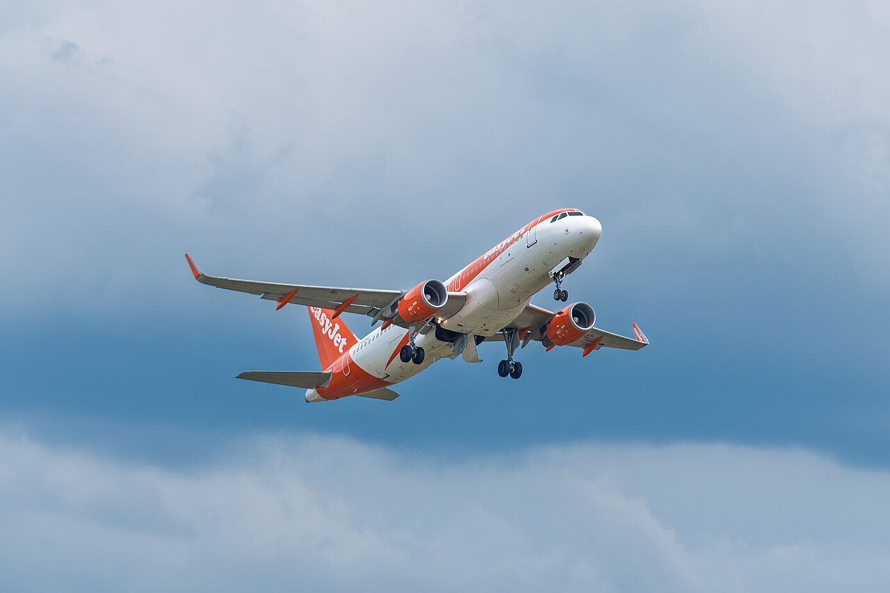 An Airbus A320-214 EasyJet aircraft in EasyJet's iconic orange and white, having just taken off from the runway. The sky is filled with wispy clouds which surround the plane. The pastel shades of the surroundings cover up distant mountains, which are only just visible.