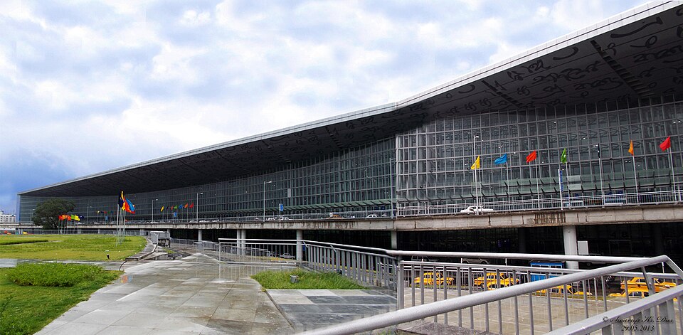 Large glass-fronted terminal with a sweeping dark roof, coloured flags along its front and yellow taxis on the road below, under a cloudy sky.