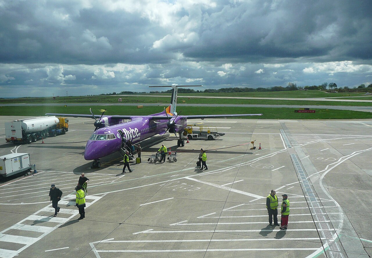 Leeds Bradford Airport, with rolling clouds that look as though a storm is brewing. The sun peeks through the thick layer of dark cloud, causing light streaks to shine downwards onto an aircraft which is stationed below, with passengers exiting.