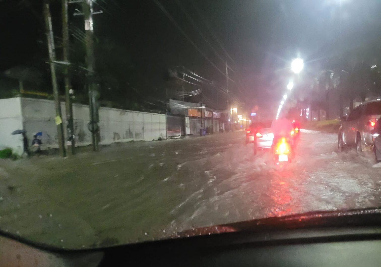 Image shows a flooded road in Pattaya, Thailand from inside a car with cars and a motorbike driving through a wave of water at night