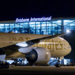 A white and gold Emirates jet parked outside of Brisbane Aiport (BNE). The jet is shown at night, and the airport terminal is illuminated by artificial lighting.