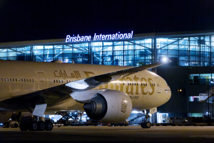 A white and gold Emirates jet parked outside of Brisbane Aiport (BNE). The jet is shown at night, and the airport terminal is illuminated by artificial lighting.