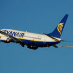 A white, blue, and yellow Ryanair jet flying in front of a clear blue sky.