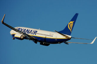 A white, blue, and yellow Ryanair jet flying in front of a clear blue sky.