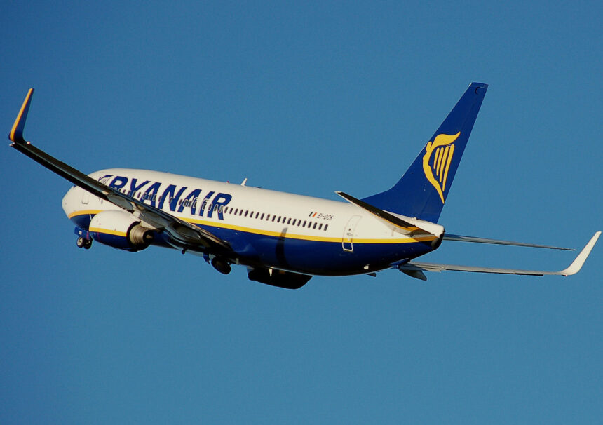 A white, blue, and yellow Ryanair jet flying in front of a clear blue sky.