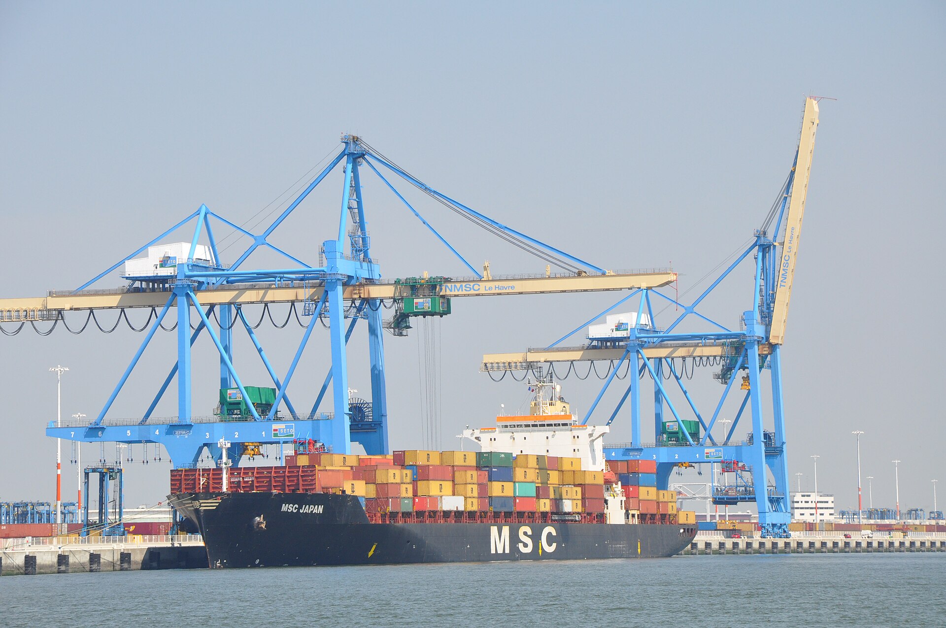 A container ship sat next to the docks at Le Havre Port in France. Blue cranes are seen behind the ship, which is loaded with containers.