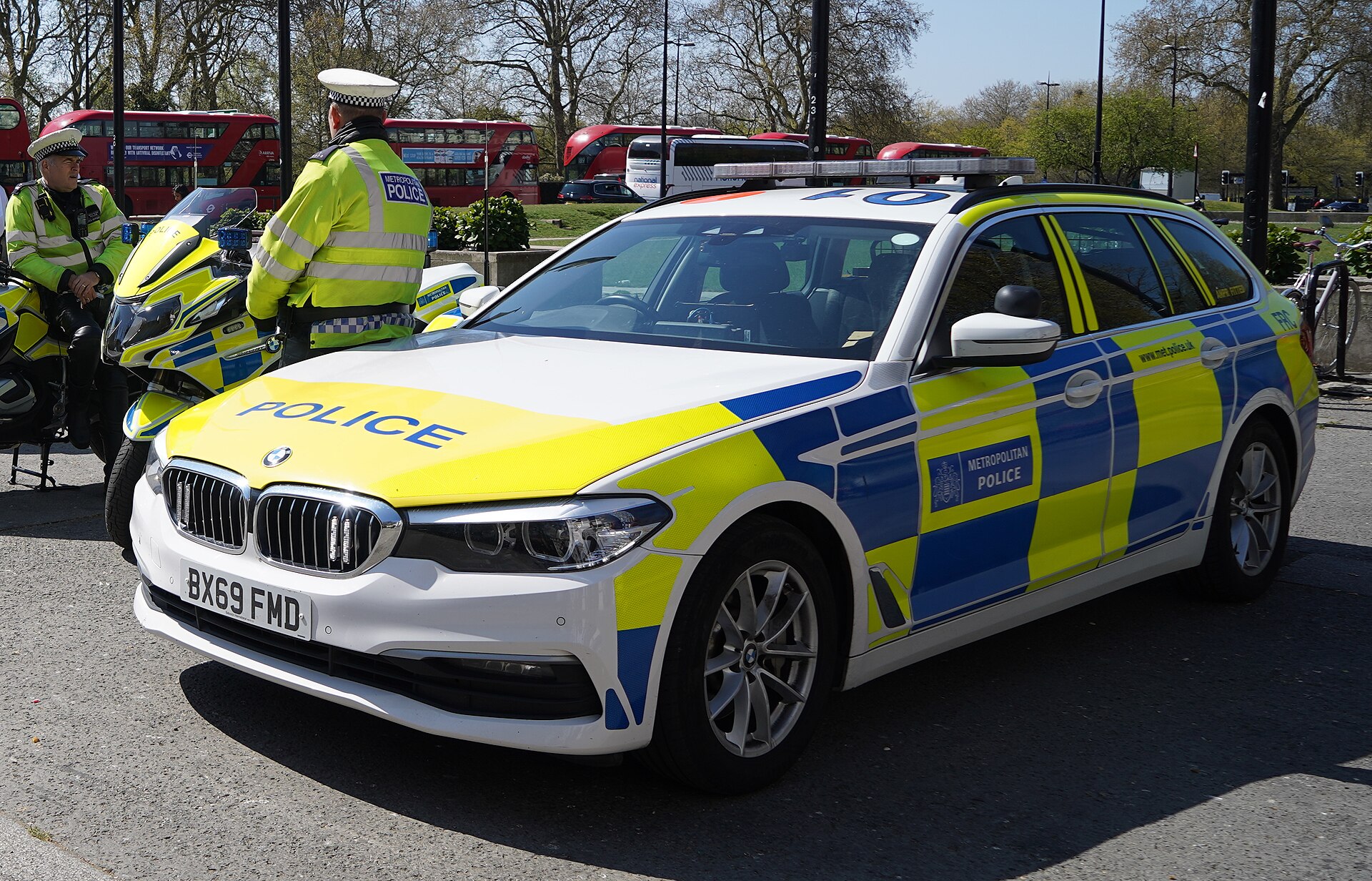 A team of police officers dressed in hi-vis uniform, stood behind a blue and green Metropolitan Police car.
