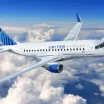 A blue and white Embraer E175 jet in the livery of United Airlines. The jet is flying over a layer of cloud.
