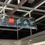 Photograph of a suspended neon sign promoting Oakland International Airport with blue “I,” red airplane symbol, and blue “OAK” lettering. Sign hangs inside a modern terminal beneath exposed beams and skylights, using bright colored icons to identify airport branding.