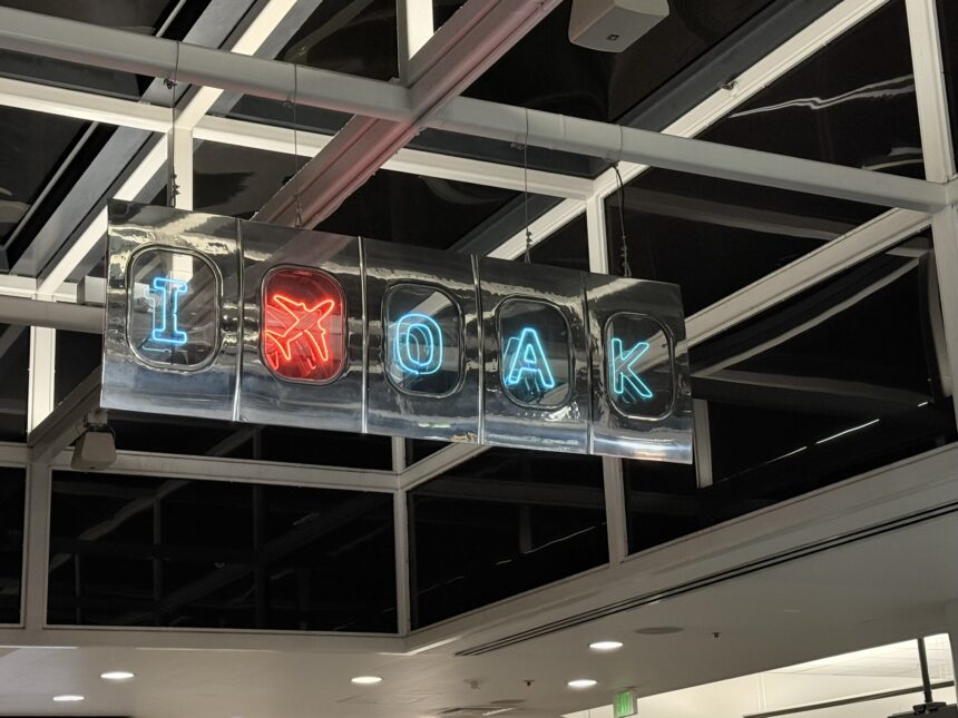 Photograph of a suspended neon sign promoting Oakland International Airport with blue “I,” red airplane symbol, and blue “OAK” lettering. Sign hangs inside a modern terminal beneath exposed beams and skylights, using bright colored icons to identify airport branding.