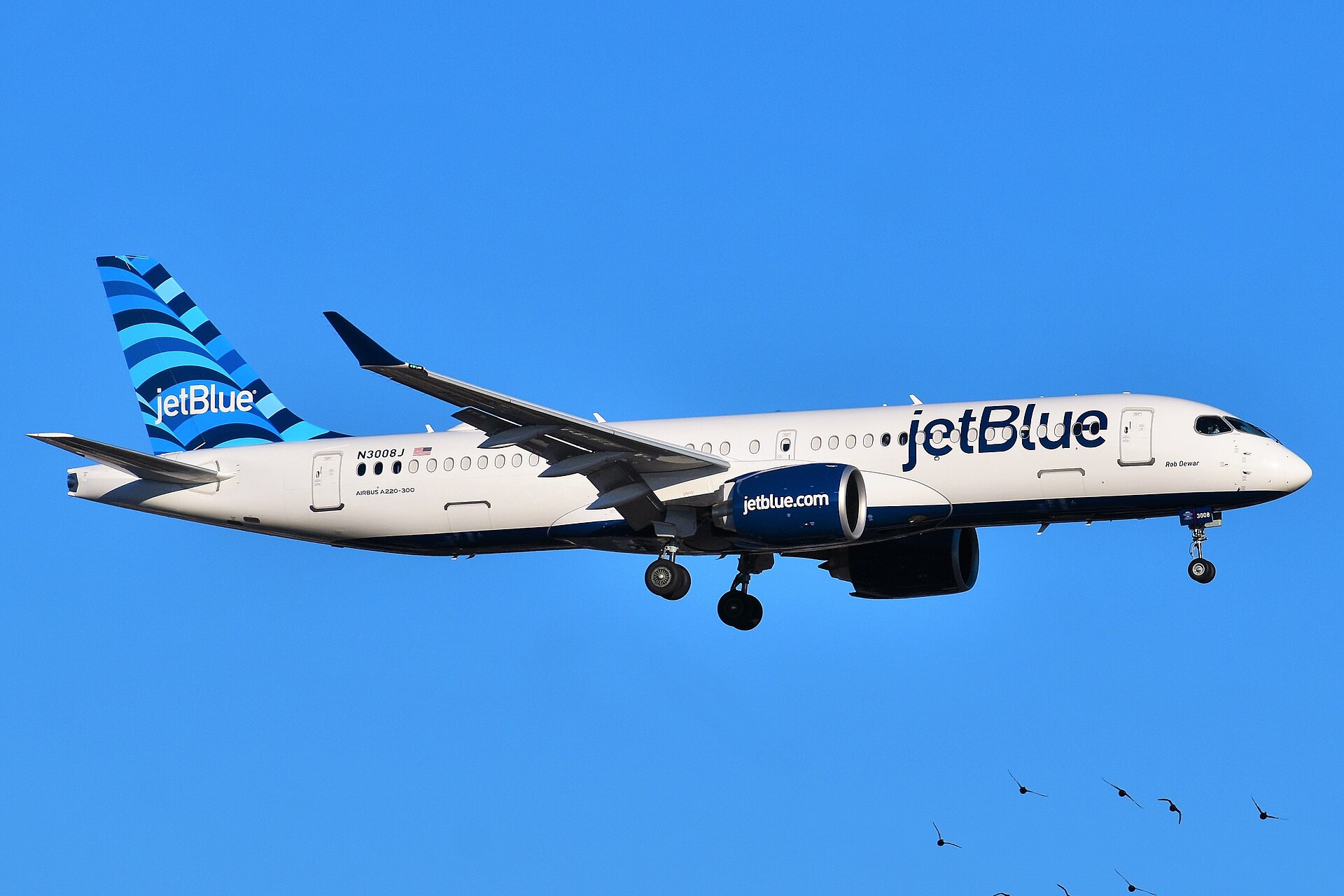 A blue and white JetBlue A220-300 flying in front of a clear blue sky.