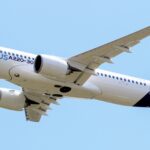 A blue and white Airbus A220-300 jet flying in front of a clear blue sky.
