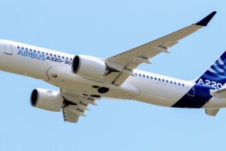 A blue and white Airbus A220-300 jet flying in front of a clear blue sky.