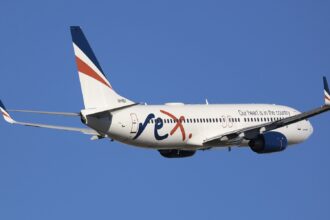A white, blue, and orange Rex Airlines jet flying in front of a clear blue sky.