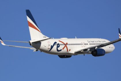A white, blue, and orange Rex Airlines jet flying in front of a clear blue sky.
