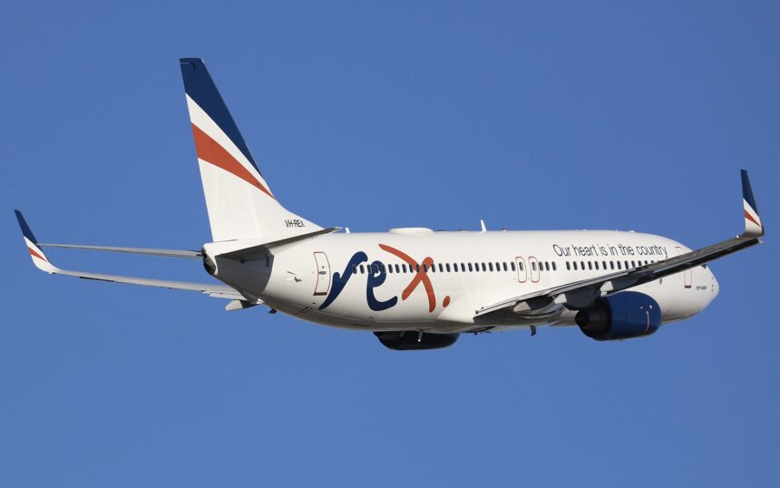 A white, blue, and orange Rex Airlines jet flying in front of a clear blue sky.