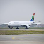 A white South African Airways Airbus jet sat on a runway. The runway is positioned behind a grassy verge, and in front of a cloudy grey sky.