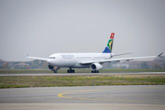 A white South African Airways Airbus jet sat on a runway. The runway is positioned behind a grassy verge, and in front of a cloudy grey sky.