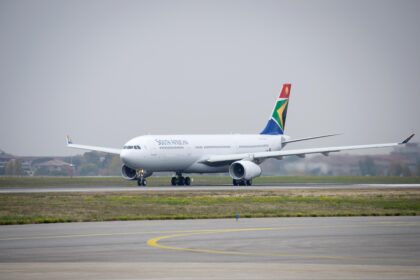 A white South African Airways Airbus jet sat on a runway. The runway is positioned behind a grassy verge, and in front of a cloudy grey sky.
