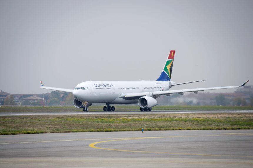 A white South African Airways Airbus jet sat on a runway. The runway is positioned behind a grassy verge, and in front of a cloudy grey sky.