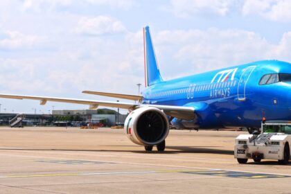 A blue ITA Airways jet sat on a runway. A small vehicle is parked next to the jet, and a cloudy sky is shown behind.