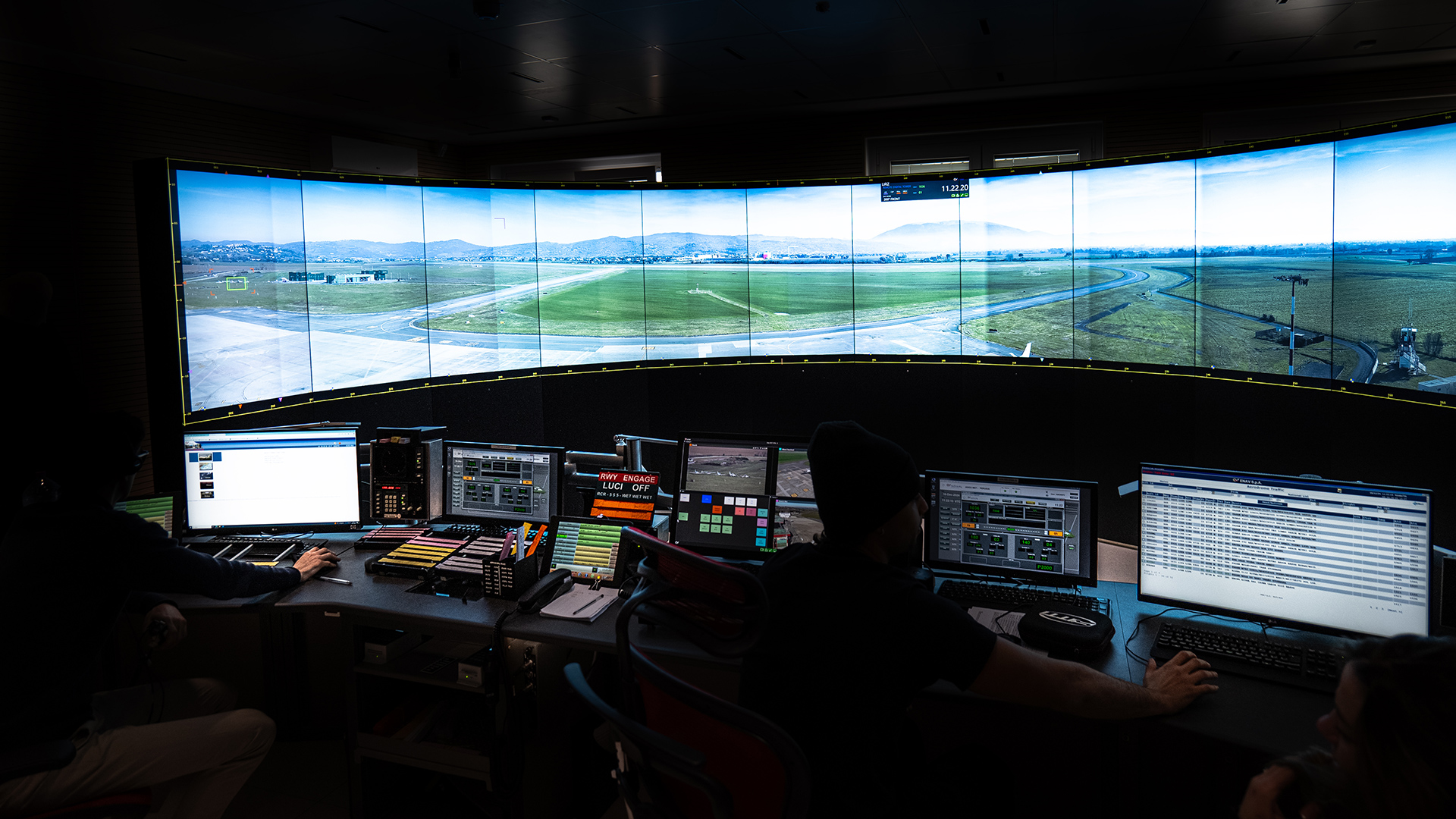 The inside of an air traffic control training booth. A large screen shows an image of an airport.