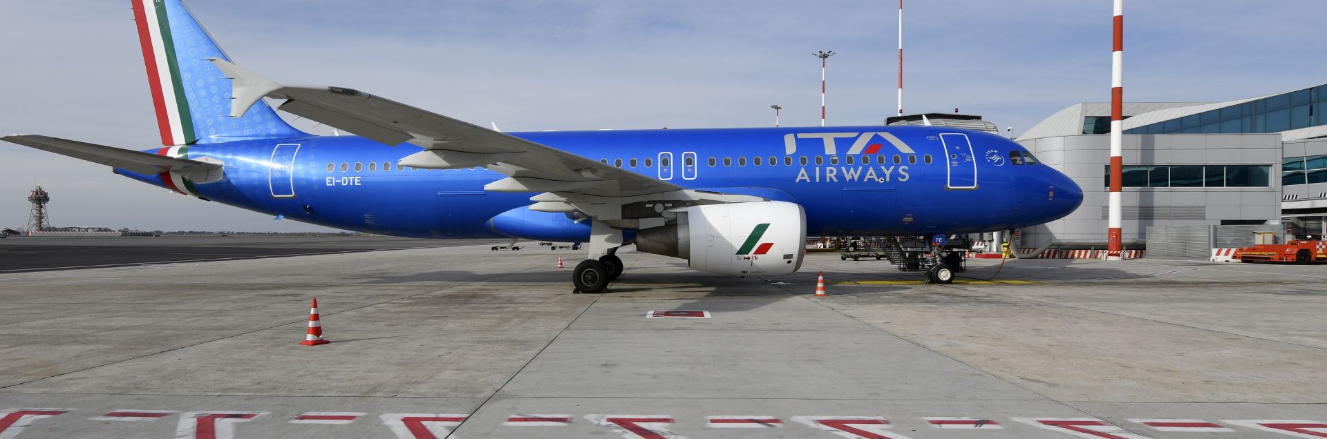 A blue ITA Airways jet parked next to the terminal at Rome Fiumicino Leonardo da Vinci International Airport (FCO). A cloudy sky is shown behind.