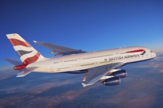 A red, white, and blue, British Airways jet flying over a layer of cloud.