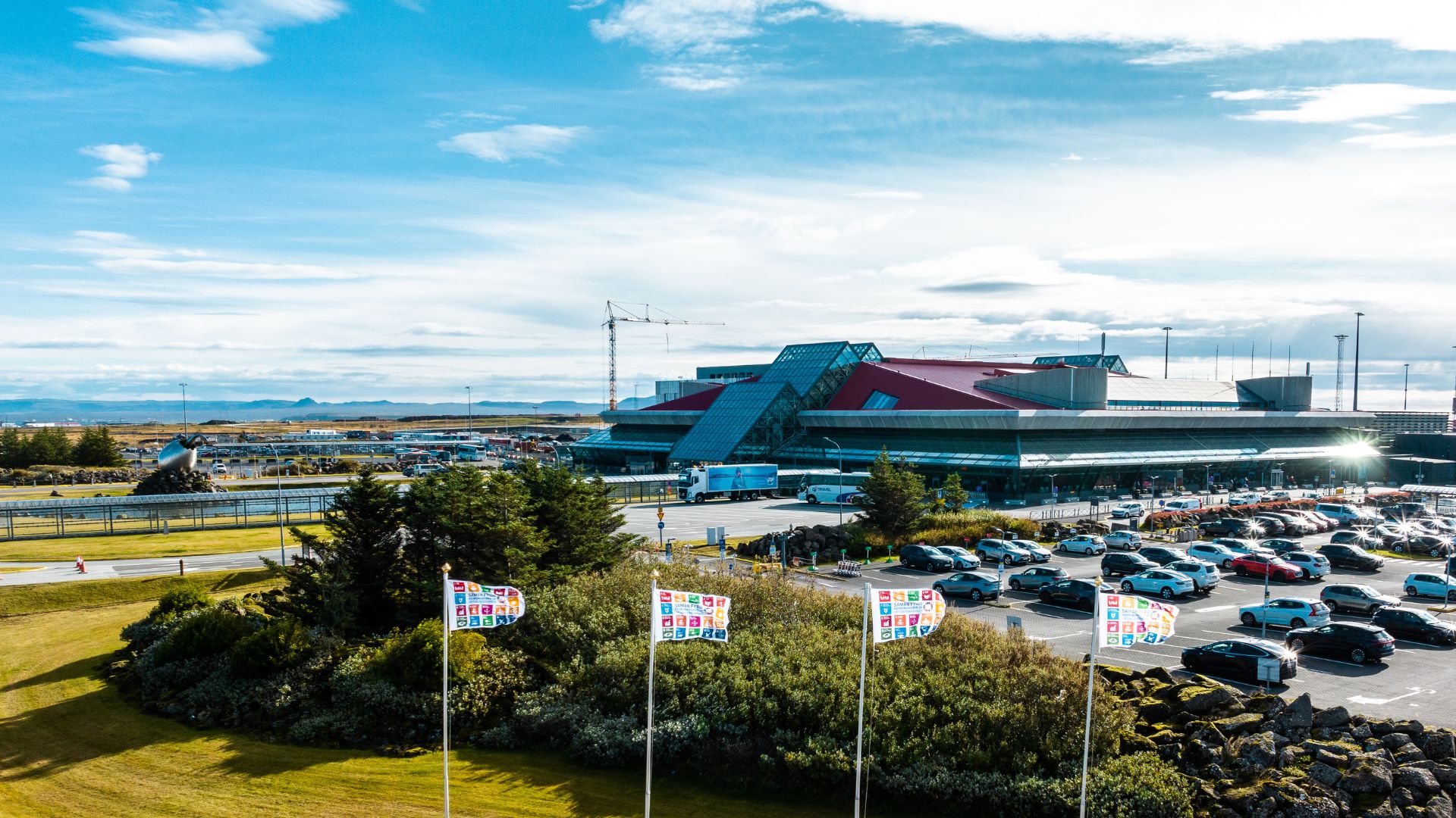 An aerial view of Keflavik International Airport (KEF) on a sunny day. The airport terminal building is visible behind a line of trees.