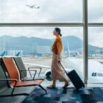 A woman in an orange jumper walks through an airport terminal pulling a suitcase.