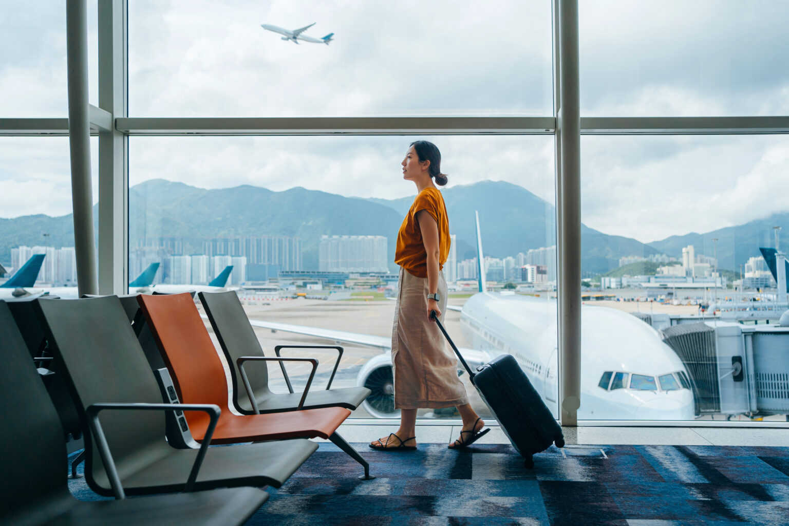A woman in an orange jumper walks through an airport terminal pulling a suitcase.