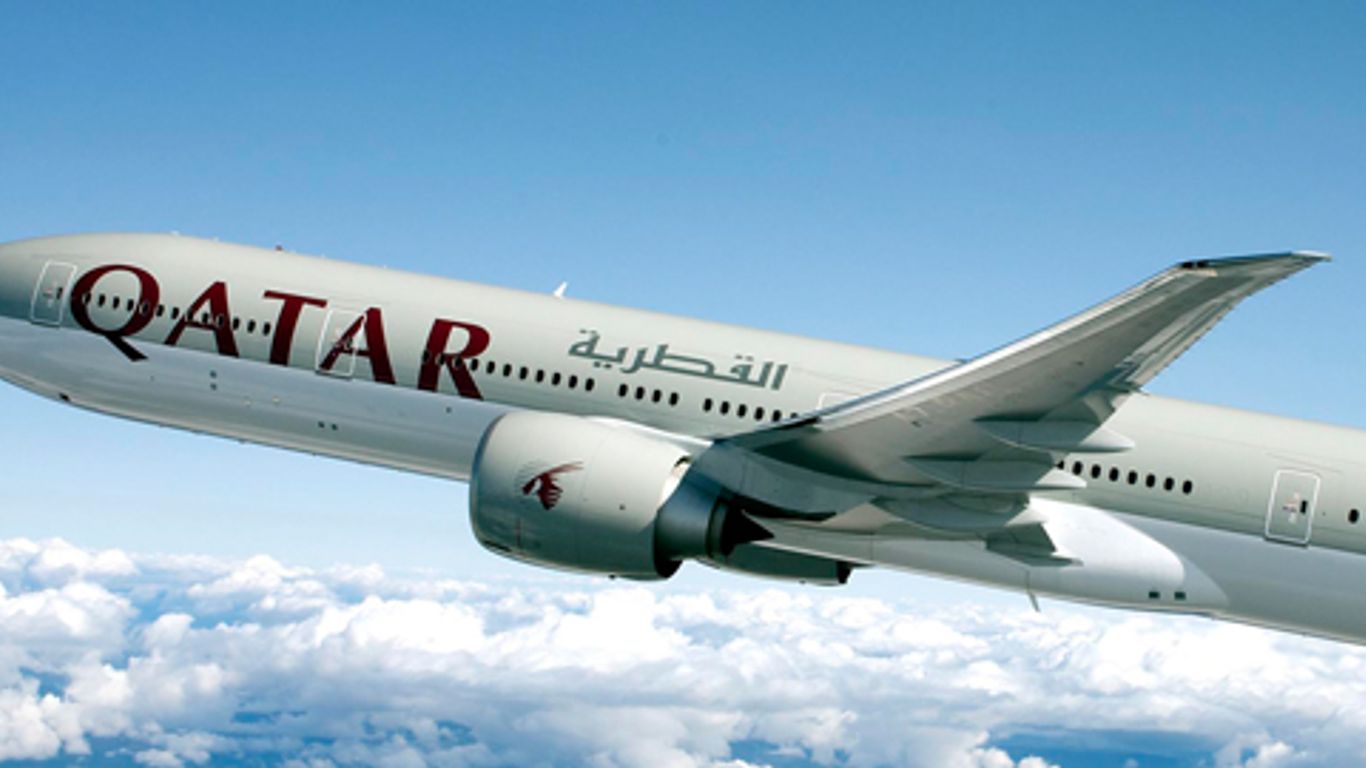 A grey, white, and purple Qatar Airways jet flying above a layer of cloud.