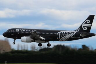 A black and white Air New Zealand jet flying above a line of dark green trees.