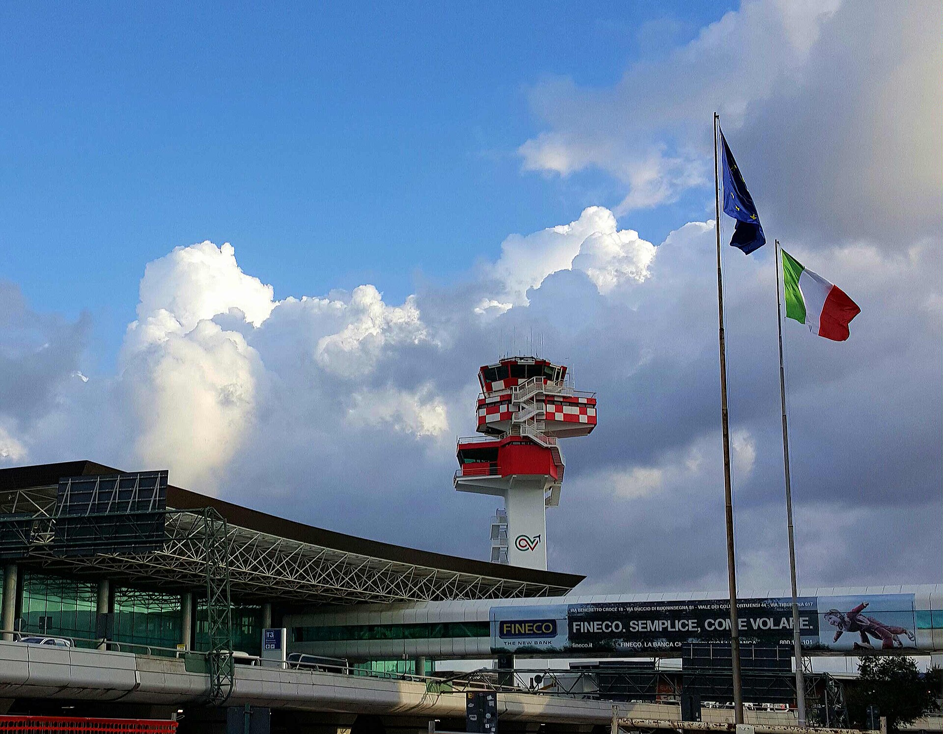 Rome Fiumicino Leonardo da Vinci International Airport (FCO). The Italian and EU flags can be seen flying in front of a cloudy sky.