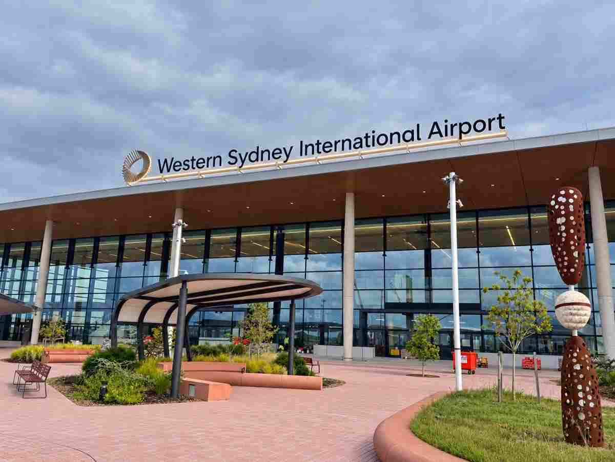 Front entrance of Western Sydney International Airport featuring a modern glass terminal building, landscaped forecourt, and dramatic cloudy sky