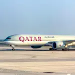 Qatar Airways Boeing 777 taxiing on a wide concrete apron under a clear, pale blue sky