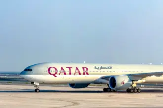 Qatar Airways Boeing 777 taxiing on a wide concrete apron under a clear, pale blue sky