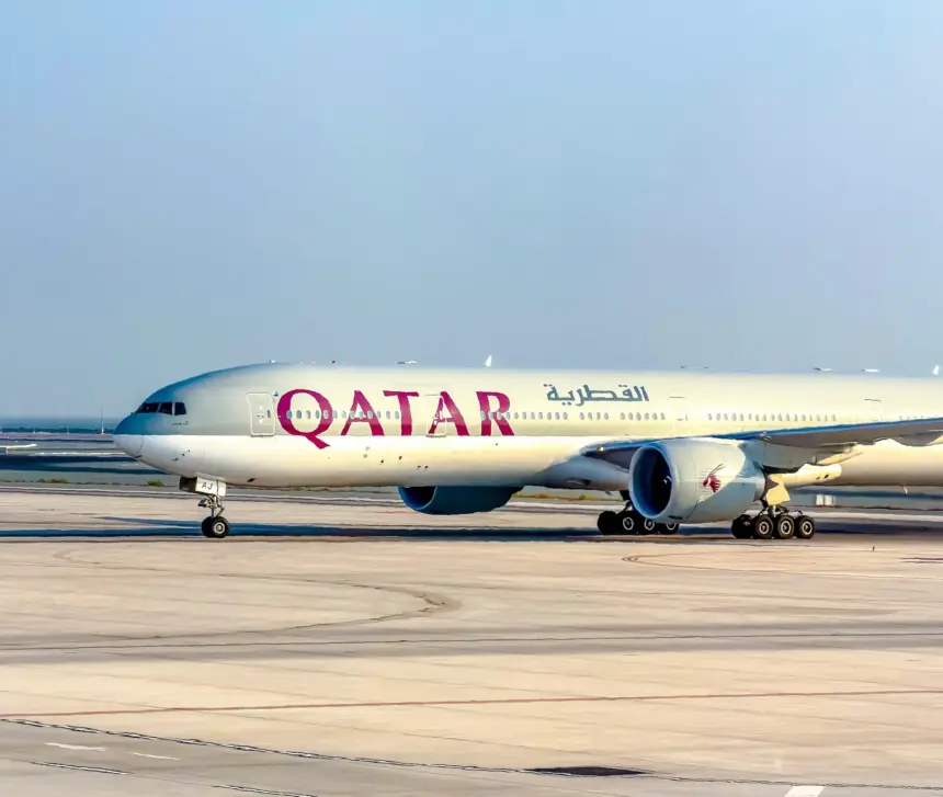 Qatar Airways Boeing 777 taxiing on a wide concrete apron under a clear, pale blue sky