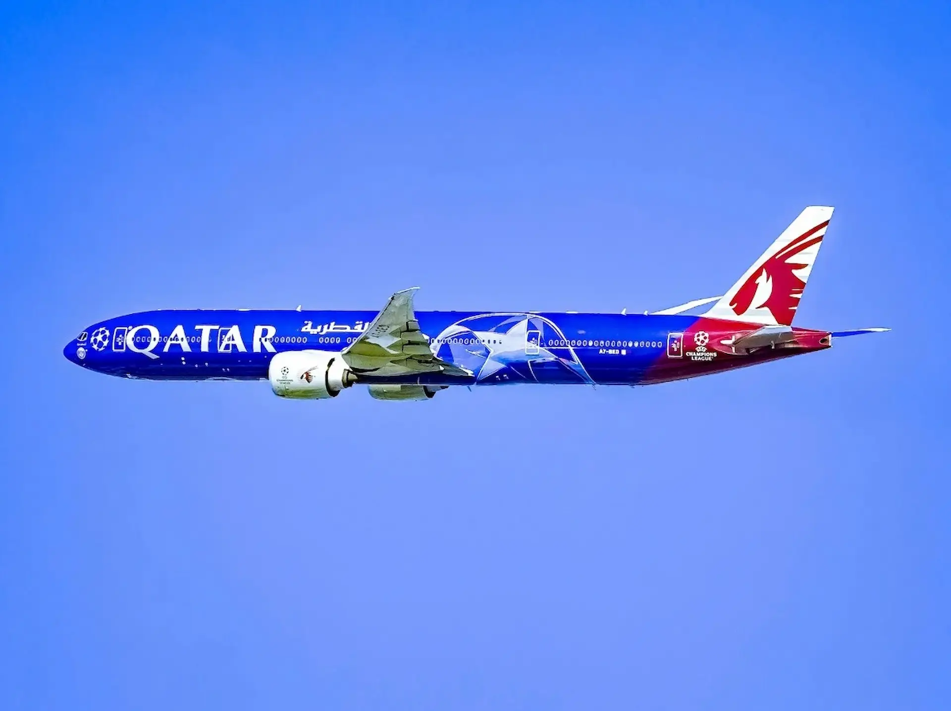 Qatar airways Boeing 777 in a blue and red UEFA Champions League livery flying against a clear blue sky, with the airline's oryx logo on the tail